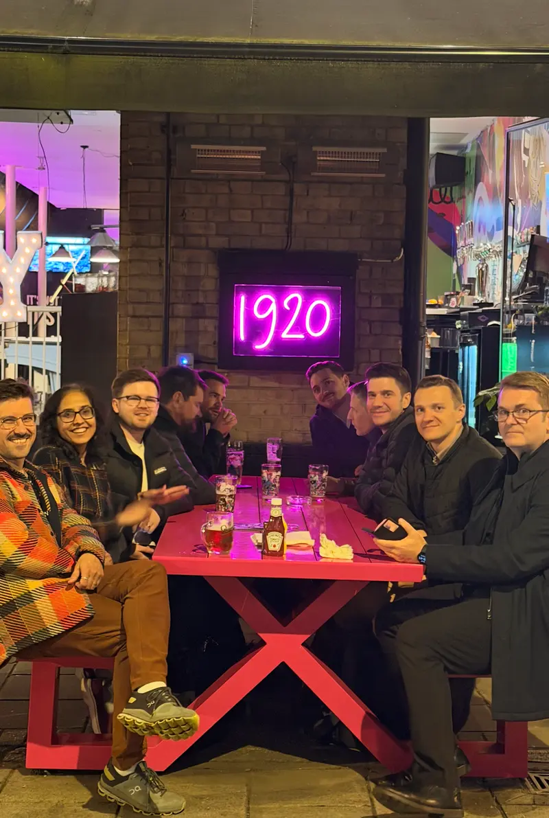 Colleagues gathered round a long table outside a bar, under a neon sign reading 1920